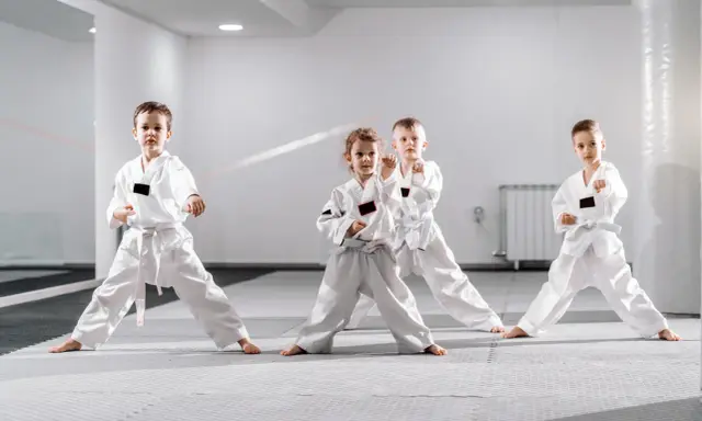 Cztery dzieci w białych kimonach ćwiczą judo dla dzieci. Stoją w pozycji bojowej na macie w jasnej sali treningowej.
