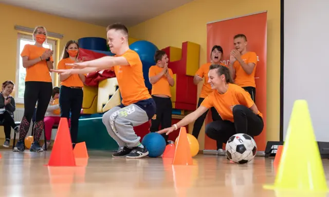 Wesołe wf dla dzieci z piłką i pachołkami. Uśmiechnięci uczestnicy i trenerzy w pomarańczowych koszulkach.
