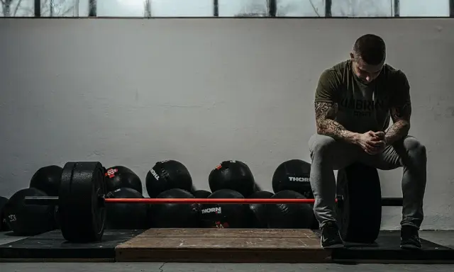 Mężczyzna z tatuażami odpoczywa po treningu crossfit w domu, otoczony piłkami lekarskimi i sztangą.