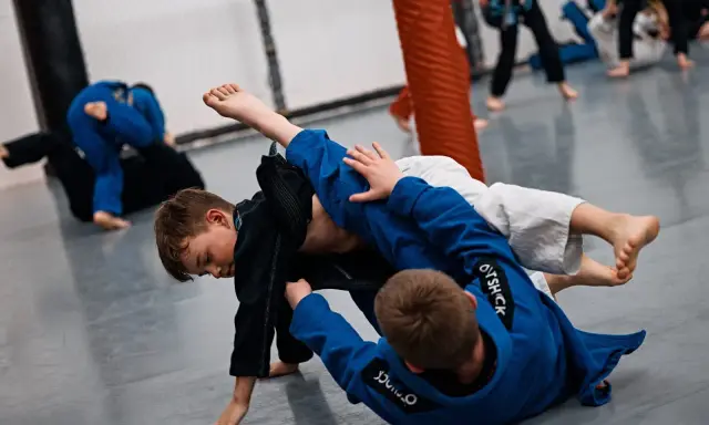 Dzieci ćwiczą jujitsu, walcząc na macie w niebieskich i czarnych kimonach.