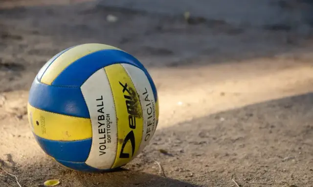 Piłka do siatkówki plażowej, niebiesko-żółta, leży na piasku. Różni się od halowej miękkością i wagą.