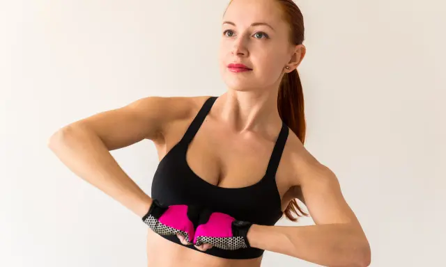 Kobieta w sportowym topie i rękawiczkach wykonuje ćwiczenia na biust, napinając mięśnie klatki piersiowej.