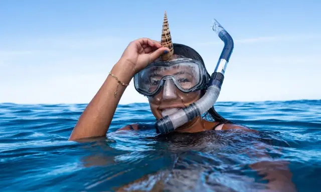 Dziewczyna w masce i fajce do snorkelingu trzyma muszlę na głowie. To właśnie snorkeling!