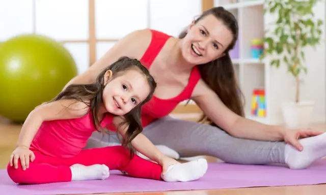 Mama i córka wykonują ćwiczenia rozciągające dla dzieci na macie. Uśmiechnięte, w różowych i szarych strojach.