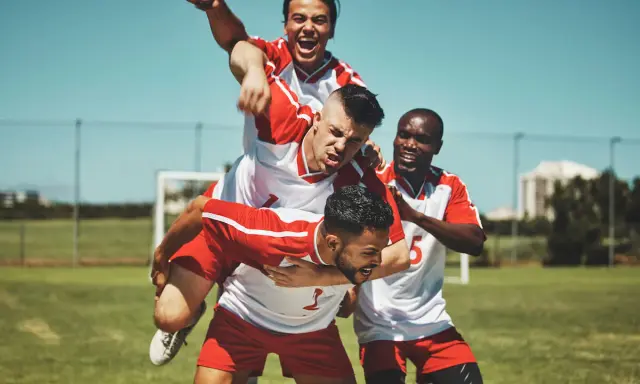 Piłkarze świętują zwycięstwo. Jeden z nich niesie kolegę na barana, krzycząc z radości. To może być celebracja hattricka, czyli zdobycia trzech bramek w jednym meczu.