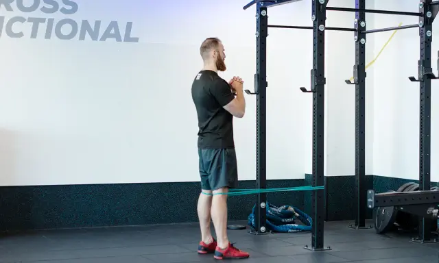 Mężczyzna wykonuje ćwiczenie spanish squat z gumą oporową wokół ud, przygotowując się do przysiadu.
