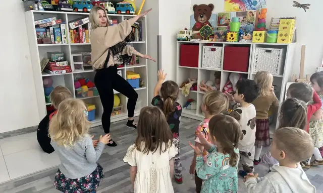 Wesołe zajęcia rytmiczne w przedszkolu. Nauczycielka tańczy z dziećmi wśród zabawek na półkach.