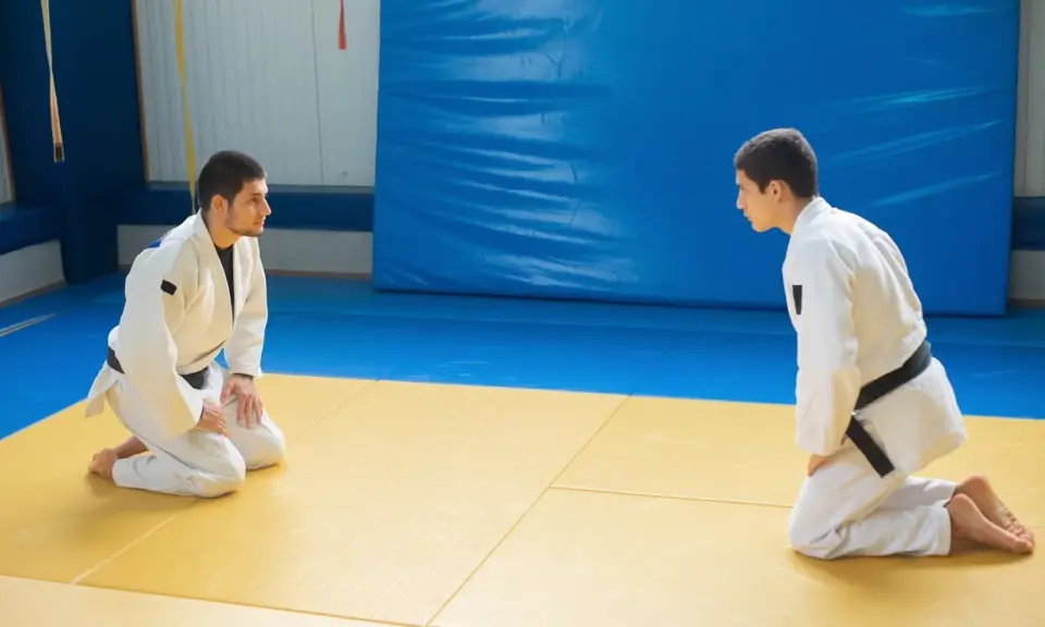 Dwóch judoków w białych kimonach, jeden z czarnym pasem judo, kłania się sobie na macie.