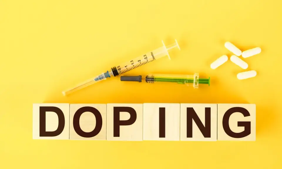 Dwa strzykawki i białe kapsułki na żółtym tle. Słowo "DOPING" napisane na drewnianych klockach.