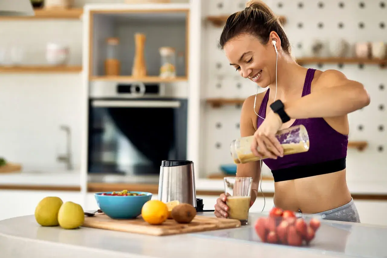 Uśmiechnięta kobieta przygotowuje smoothie, idealne jako co zjesc przed biegiem. Na blacie owoce i blender.
