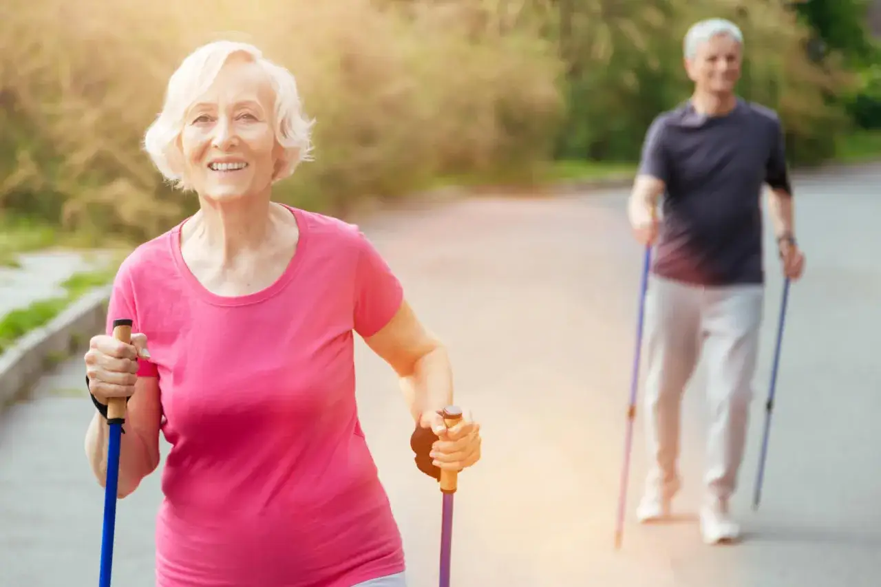 Uśmiechnięta starsza pani i pan w tle uprawiają nordic walking, spalając kalorie na świeżym powietrzu.
