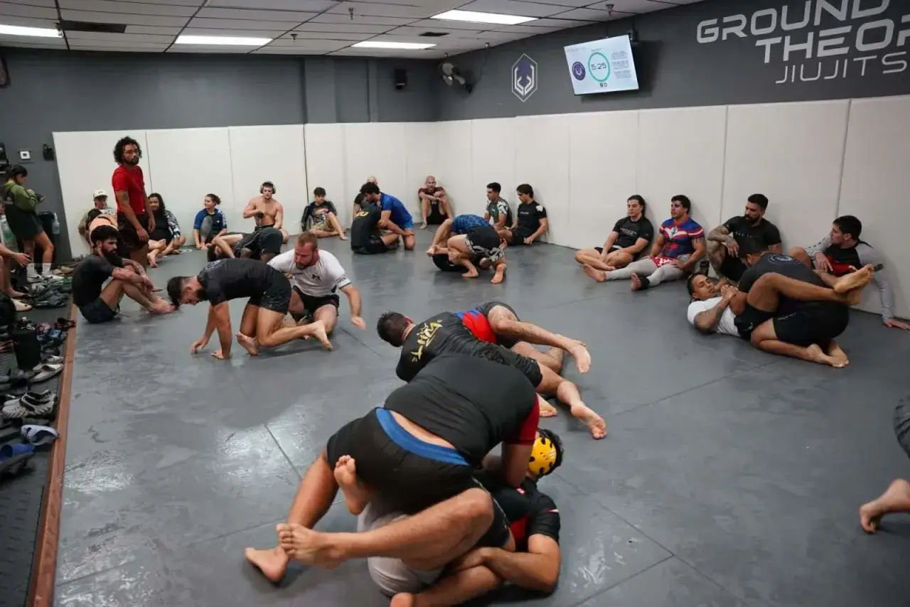 Grupa ćwiczy no gi bjj na macie. Niektórzy siedzą, inni sparują.