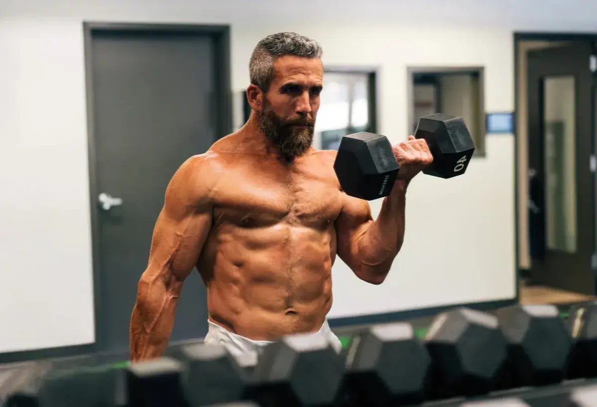 Mężczyzna z brodą ćwiczy biceps z hantlem, osiągając imponującą rekompozycję sylwetki.