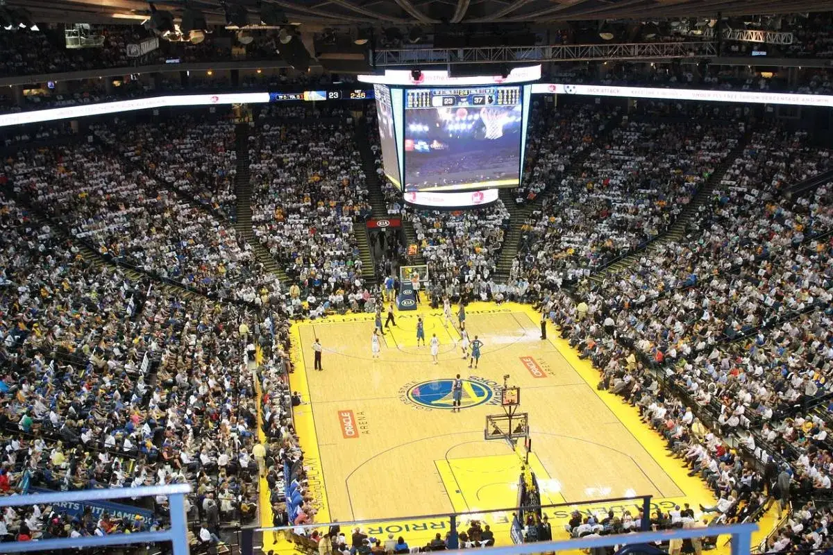 Mecz koszyk&oacute;wki w Oracle Arena. Na boisku gracze, trybuny wypełnione kibicami. Na telebimie widać wynik, kt&oacute;ry sugeruje, że gra jest w końcowej fazie, być może ostatni z czterech kwart w koszyk&oacute;wce.