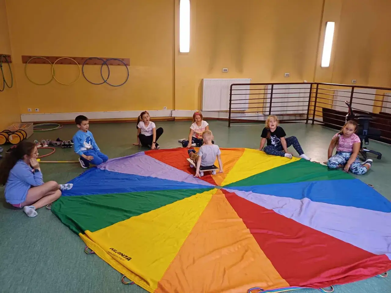 Dzieci siedzą na tęczowym spadochronie, a jeden chłopiec zjeżdża na deskorolce. Wesołe zajęcia dodatkowe dla 4 latka.