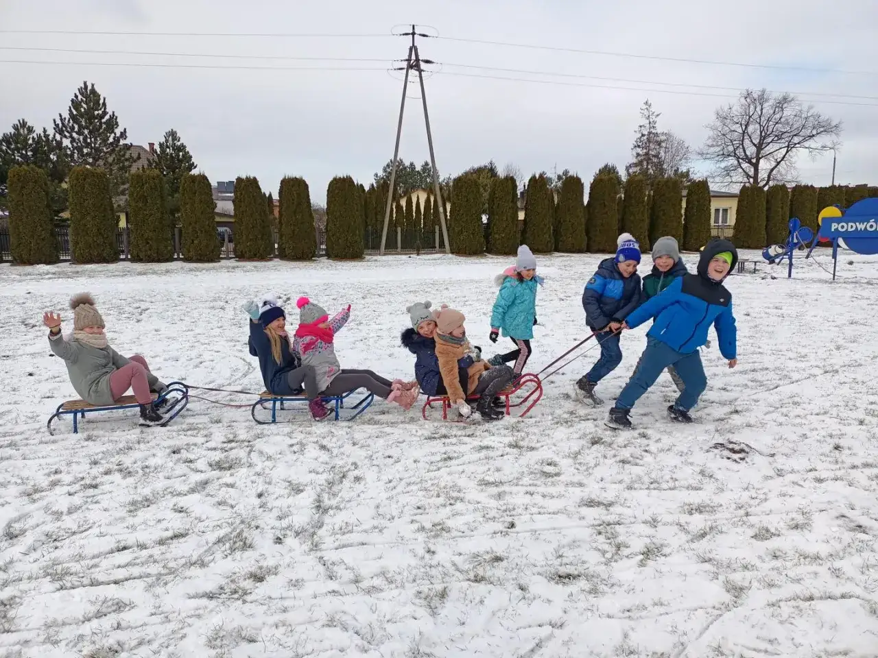 Radosne zimowe zabawy dla dzieci na sankach. Grupa dzieci ciągnie się nawzajem po śniegu, śmiejąc się i ciesząc zimową aurą.