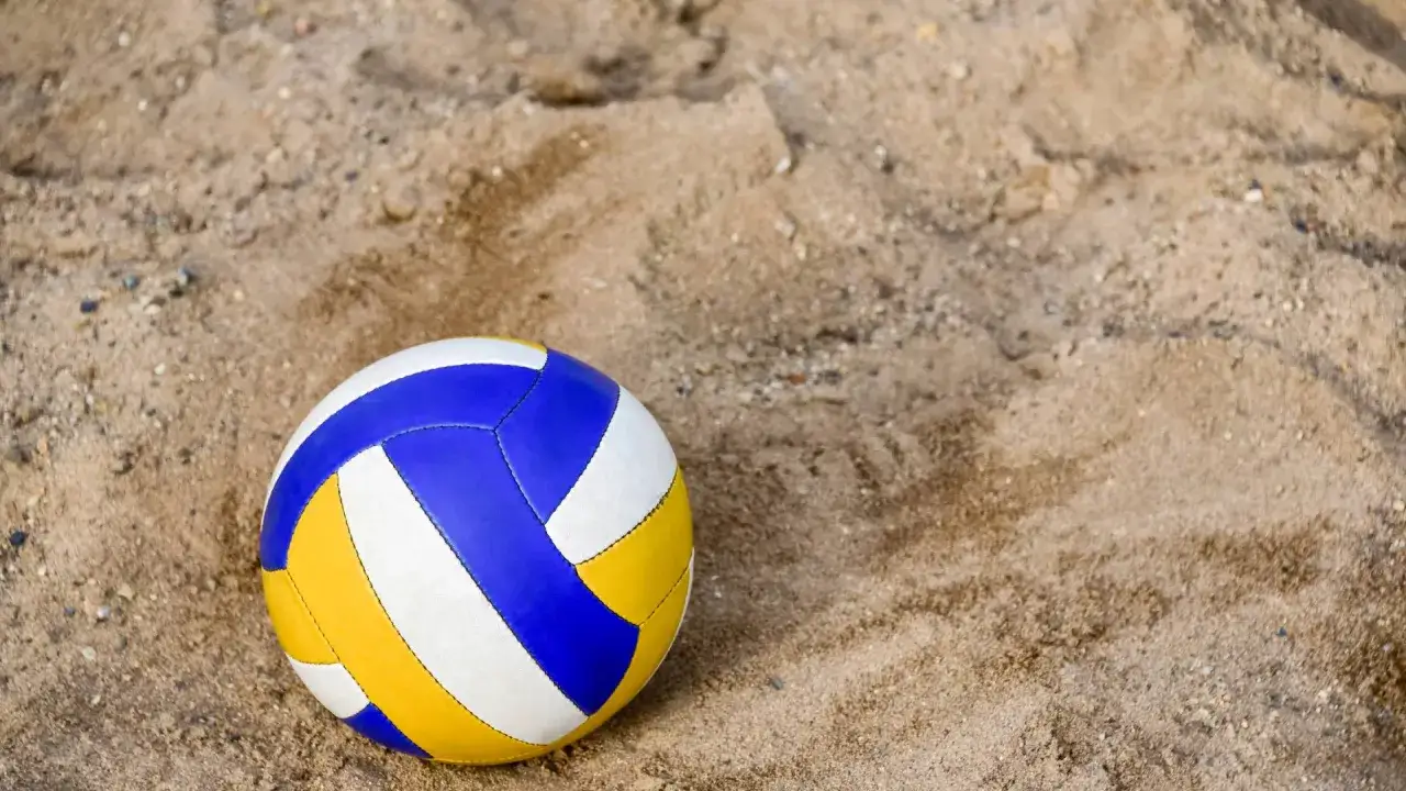 Piłka do siatkówki plażowej, żółto-biało-niebieska, leży na piaszczystej plaży. Różni się od halowej miękkością i kolorem.
