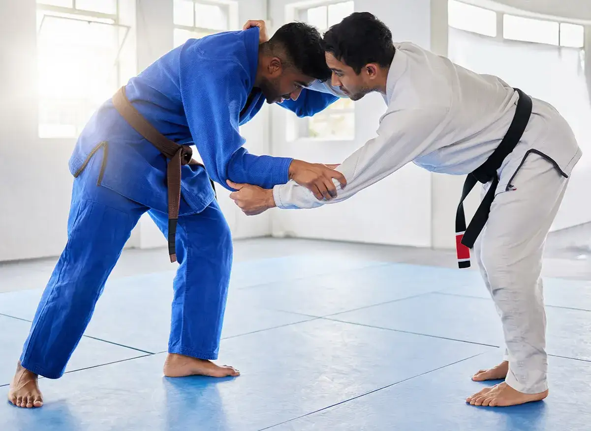Dwóch mężczyzn w kimonach do BJJ, jeden w niebieskim z brązowym pasem, drugi w białym z czarnym pasem, ćwiczy chwyty.