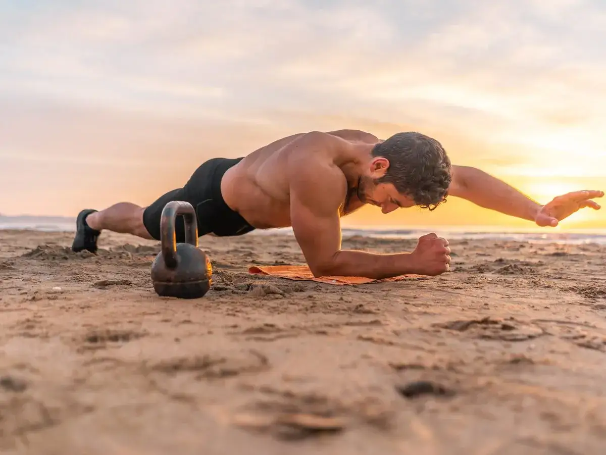 Mężczyzna na plaży wykonuje ćwiczenie w desce, obok kettlebell. Cel: 300 brzuszków.