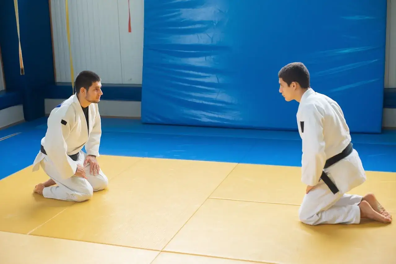 Dwóch judoków w białych kimonach, jeden z czarnym pasem judo, kłania się sobie na macie.