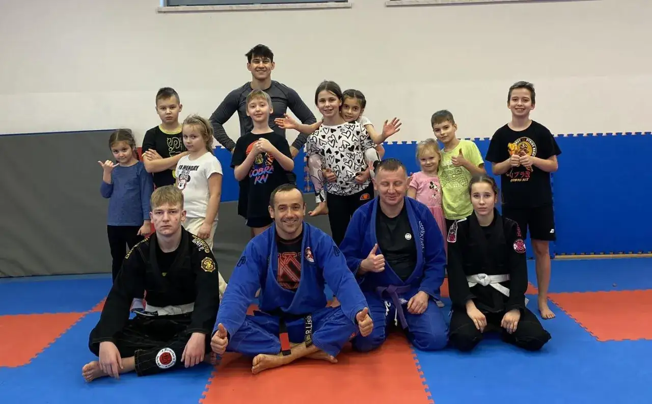 Grupa dzieci i trenerów na macie po treningu mma dla dzieci. Wszyscy uśmiechnięci, niektórzy pokazują kciuki w górę.