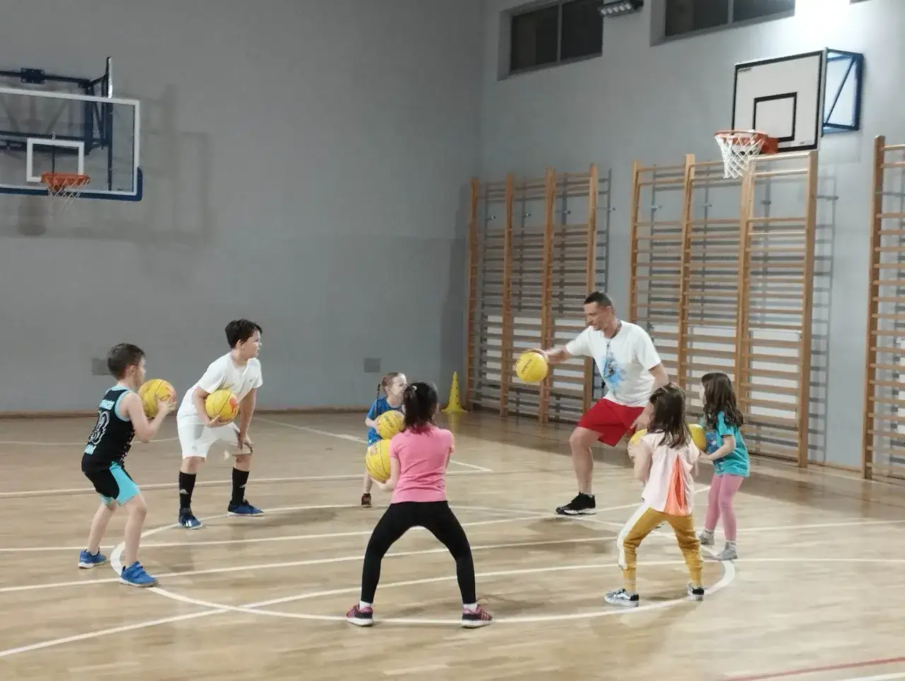 Trener prowadzi zajęcia z koszyk&oacute;wki dla dzieci. Mali gracze ćwiczą rzuty i podania na sali gimnastycznej.