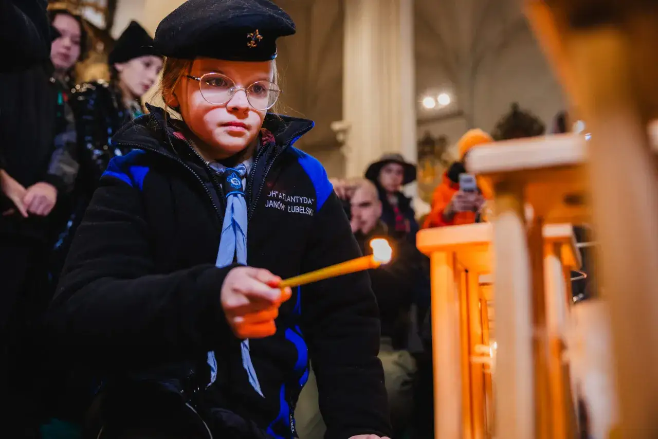 Dziewczynka w harcerskim mundurku i beretce zapala świecę. W tle widać innych uczestników, być może podczas gry harcerskiej w pomieszczeniu.