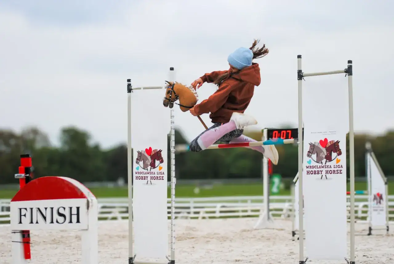 Dziewczynka w brązowej bluzie i niebieskiej czapce skacze przez przeszkodę na kucyku. To emocjonujące hobby horsing!