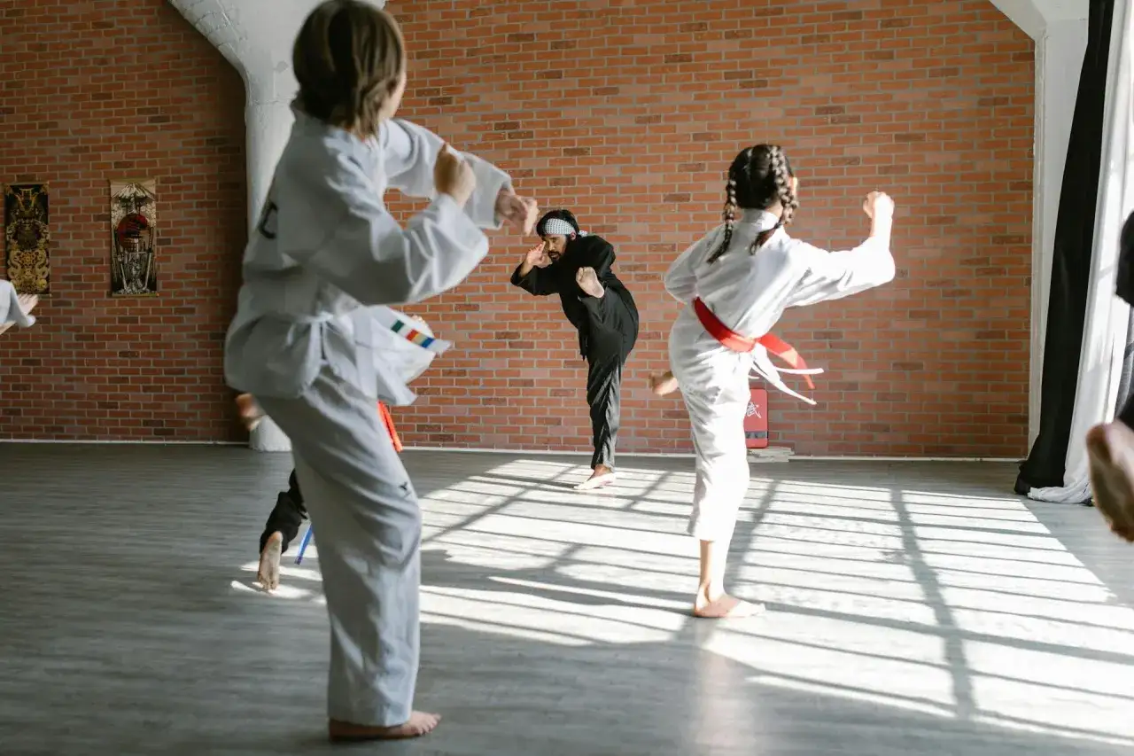 Dzieci ćwiczą judo, wykonując kopnięcia. Trener w czarnym stroju demonstruje technikę.