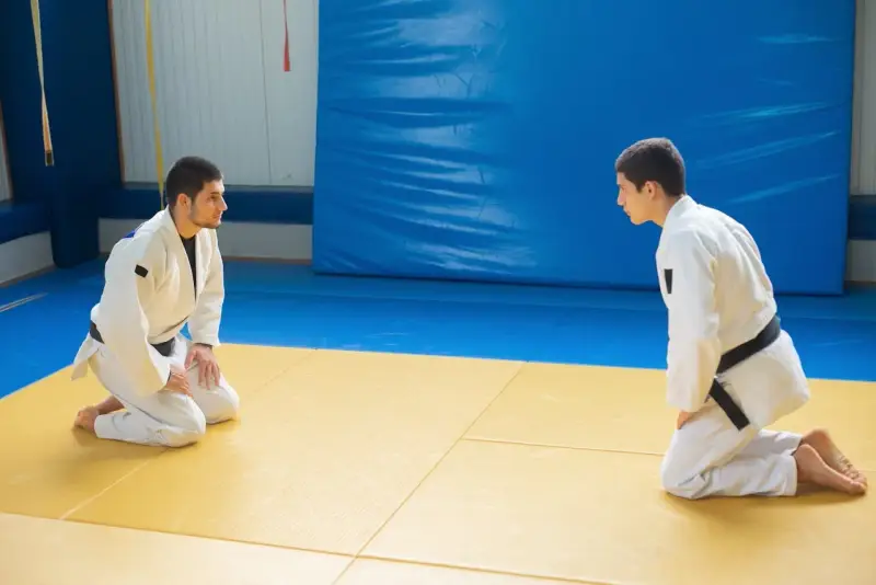 Dwóch judoków w białych kimonach, jeden z czarnym pasem judo, kłania się sobie na macie.
